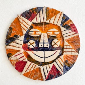 May include: A ceramic sun face with a smiling expression. The sun has orange, red, blue, and white accents. The sun's rays are triangular and radiate from the center. The face has a nose, eyes, and a wide smile.