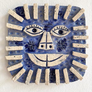 Puede incluir: Escultura de rostro de cerámica azul con expresión sonriente. El rostro tiene ojos blancos, nariz blanca y boca ancha y curvada. Piezas rectangulares blancas forman un patrón de rayos de sol alrededor del rostro. Dos diseños azules en forma de estrella están en el rostro.