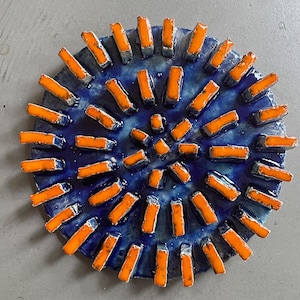 May include: A circular ceramic art piece. The base is a deep blue with a textured finish. Orange rectangular tiles are arranged in a radial pattern, creating a sunburst or floral design. The piece is likely handmade and would suit a modern home.