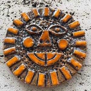 Puede incluir: Una placa de pared circular de cerámica que presenta un rostro de sol estilizado con un esmalte naranja brillante. El sol tiene un fondo marrón oscuro texturizado, rayos naranjas prominentes, dos ojos almendrados, una nariz triangular y una amplia sonrisa.