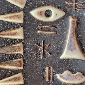 Puede incluir: Una superficie de cerámica gris oscuro moteada con formas de arcilla clara en relieve, que incluyen un ojo, un asterisco en forma de estrella, una forma triangular alta, un par de labios y varias barras y triángulos geométricos.