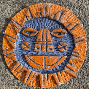 May include: A circular ceramic wall plaque featuring a stylised sun face with a textured orange and deep blue glaze. The design includes radiating triangular sunbeams, almond-shaped eyes, a rectangular nose, and a wide, smiling mouth.
