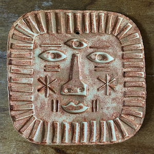 May include: A square ceramic wall plaque with a textured, speckled terracotta and cream finish. The surface features a stylised face with a third eye, two almond-shaped eyes, a triangular nose, a small mouth, and geometric star and line symbols.