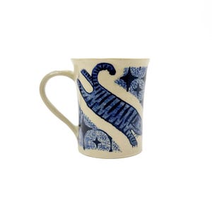 Könnte beinhalten: Cremefarbene Keramik-Tasse mit blauem, abstraktem Design. Die Tasse hat einen gebogenen Henkel und ist mit einer blau gestreiften Form und Sternmustern verziert. Das Design wiederholt sich um die Tasse.