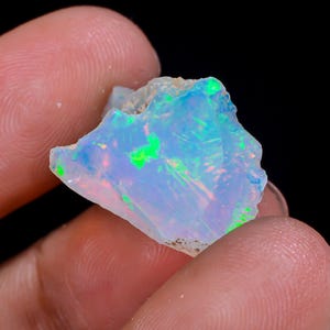 Può includere: Una pietra di opale grezza, iridescente, con una miscela di tonalità blu, verdi e rosa. La struttura cristallina della pietra è visibile. È tenuta da una mano su sfondo nero. L'opale è un minerale naturale.