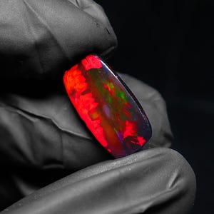 May include: A rectangular black opal gemstone with vibrant red, green, and purple internal fire. The polished stone is held against a black background, showcasing its color play. The opal's dimensions are approximately 1.5 inches long.