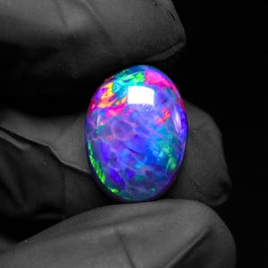 May include: An oval-shaped opal gemstone with a vibrant display of colors. The stone features a mix of blues, greens, pinks, and purples, creating a dynamic and eye-catching appearance. The opal is held against a black background.