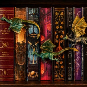 Peut inclure: Trois petites figurines de dragon détaillées dans des tons vert et or sont placées sur une rangée de livres reliés. Les dragons présentent des écailles texturées, des ailes déployées et de longues queues, semblant ramper sur les dos.