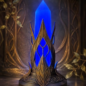 Pode incluir: Uma ponta de cristal azul brilhante inserida numa base de metal intrincada de cor bronze escuro. A base apresenta estruturas verticais pontiagudas em forma de folha e padrões de filigrana que rodeiam a base da pedra azul iluminada.