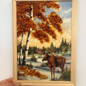 Op de afbeelding: Ingelijst kunstwerk met een eland in een besneeuwd landschap met berken en herfstbladeren. Het kunstwerk heeft een licht houten frame en een gedetailleerde scène met warme, aardse tinten.