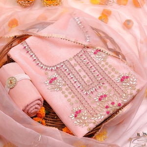 Può includere: Un completo salwar kameez rosa chiaro ricamato con dettagli oro e rossi. Il set include un top, un fondo e una sciarpa, disposti in un cestino di vimini con accenti floreali. È incluso anche un braccialetto abbinato.