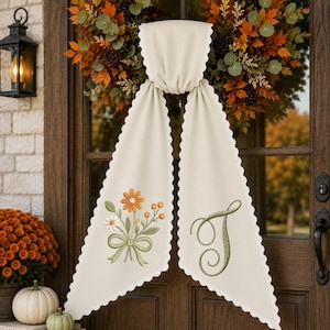 May include: A cream-coloured fabric door hanger with scalloped edges, featuring an embroidered bouquet of orange and white flowers on the left side and a large, decorative green embroidered letter T on the right side, draped over a fall wreath.