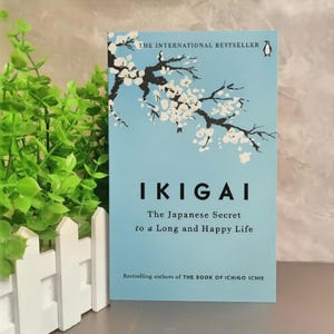 May include: A book titled "IKIGAI" with a light blue cover. The cover features a branch with white flowers and the text "The Japanese Secret to a Long and Happy Life." The book is a bestseller.