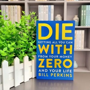 May include: A hardcover book titled "DIE" in large yellow letters on a blue background. The book's subtitle reads "Getting All You Can With From Your Money Zero And Your Life" by Bill Perkins. The book is displayed in front of a white picket fence and green foliage.