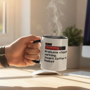 Op de afbeelding: Witte koffiemok met een zwart handvat en rand, met de tekst "Brain.exe stopped working. Insert Coffee to Reboot." Stoom stijgt op uit de mok. De mok wordt door een persoon vastgehouden.