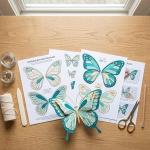 May include: A printable butterfly template on white paper featuring teal and cream wing designs, a three-dimensional paper butterfly model, a spool of white twine, a bone folder, a pair of metal scissors, and a wooden pencil on a wood surface.