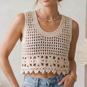 Può includere: Un top all'uncinetto senza maniche color crema, con scollo tondo e dettagli floreali lungo l'orlo. Il design a trama aperta è visibile su tutto il top. La modella indossa jeans blu e braccialetti dorati.