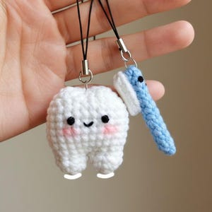 May include: Crocheted tooth and toothbrush charms. The tooth is white with a smiling face and pink cheeks. The toothbrush is blue with a white bristle area. Both charms have black cords and silver-colored metal attachments.