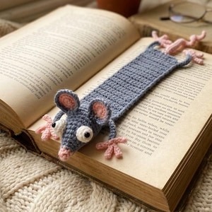 May include: A crocheted bookmark shaped like a gray mouse with pink ears and feet, lying on the pages of an open book. The bookmark is handmade and adds a whimsical touch to reading. The book is open to a page of text.
