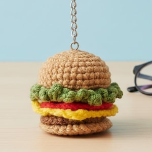 Op de afbeelding: Een gehaakte hamburger sleutelhanger met een zilveren ketting en ring. De burger heeft een bruin broodje, groene sla, rode tomaat, gele kaas en een bruine patty. De achtergrond is een lichtblauwe muur en een houten tafel.