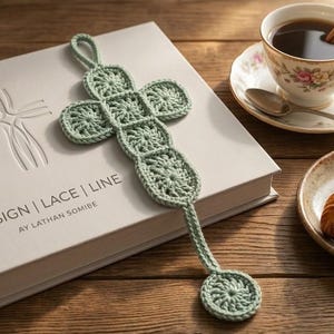 May include: A light green crocheted cross-shaped bookmark rests on a book with the text "DESIGN | LACE | LINE". A cup of coffee with a cinnamon stick and a croissant are also visible.