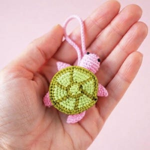 May include: A small, handmade crochet turtle keychain featuring a light pink head, limbs, and tail, with a textured, multi-toned lime and olive green shell. The turtle includes a matching pink yarn loop attached to the top of its head for hanging.