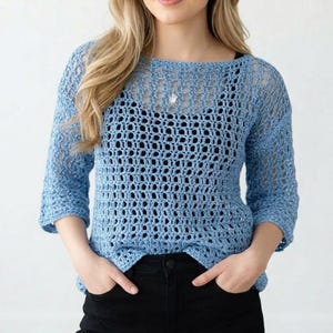 May include: A light blue crochet sweater with a loose, open weave. The sweater has three-quarter sleeves and a boat neck. The model is wearing black pants. The sweater is a stylish, handmade garment.