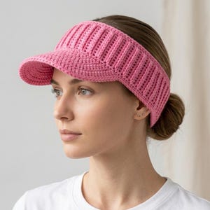 以下が含まれることがあります： ピンク色のリブ編みデザインの、かぎ針編みバイザー。バイザーは、湾曲したつばと、頭の周りにフィットする幅広のバンドが付いています。帽子は柔らかく、質感のある糸で作られています。バイザーは、日焼け防止のためのスタイリッシュなアクセサリーです。
