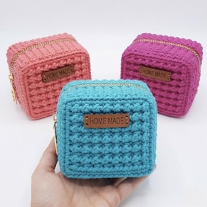 May include: Three square crochet pouches in pink, magenta, and teal. Each pouch features a gold-tone zipper closure and a rectangular brown leather-like tag centered on the front with the text "HOME MADE" embossed in a dark brown font.