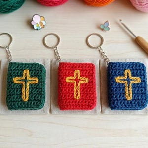 Op de afbeelding: Drie gehaakte boeksleutelhangers in groen, rood en blauw, elk met een gouden kruisontwerp. De sleutelhangers zijn bevestigd aan zilveren kettingen en liggen op kleine, beige stoffen vierkantjes. Kleurrijke bollen wol en een haaknaald zijn op de achtergrond.
