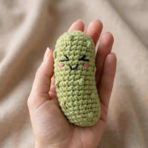 Puede incluir: Un pequeño pepinillo verde tejido a ganchillo con una cara sonriente, con ojos y boca bordados en negro y mejillas rosas. El juguete de peluche hecho a mano se sostiene en una mano, mostrando su textura detallada y expresión alegre.