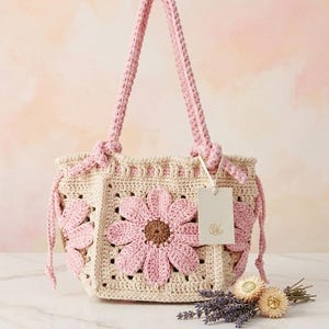 Könnte beinhalten: Eine cremefarbene Häkeltasche mit rosa Gänseblümchen-Applikationen. Die Tasche hat rosa Griffe und Kordelzüge mit einer passenden rosa Gänseblümchen auf der Vorderseite. Ein kleines Etikett hängt am Griff. Die Tasche ist handgefertigt.