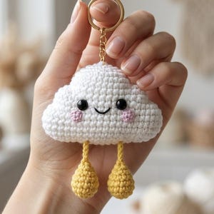 Op de afbeelding: Een witte gehaakte wolken-sleutelhanger met twee zwarte kraaloogjes, een geborduurde zwarte glimlach en twee roze geborduurde wangen. Twee gele gehaakte druppelvormen hangen onderaan de wolk aan korte gele koordjes aan een gouden ring.