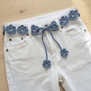 May include: A pair of white denim jeans featuring a light blue crocheted belt. The belt is constructed from a series of interconnected floral motifs and includes two long, dangling ties that each end in a single matching crocheted flower.
