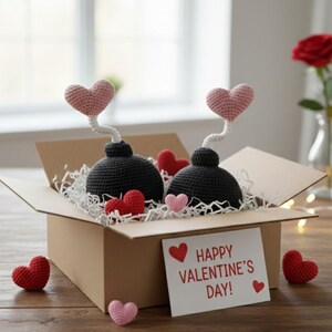 May include: A cardboard box filled with crochet Valentine's Day decorations. Two black crochet bombs with pink heart tops sit among red and pink crochet hearts. A card reads "Happy Valentine's Day!"