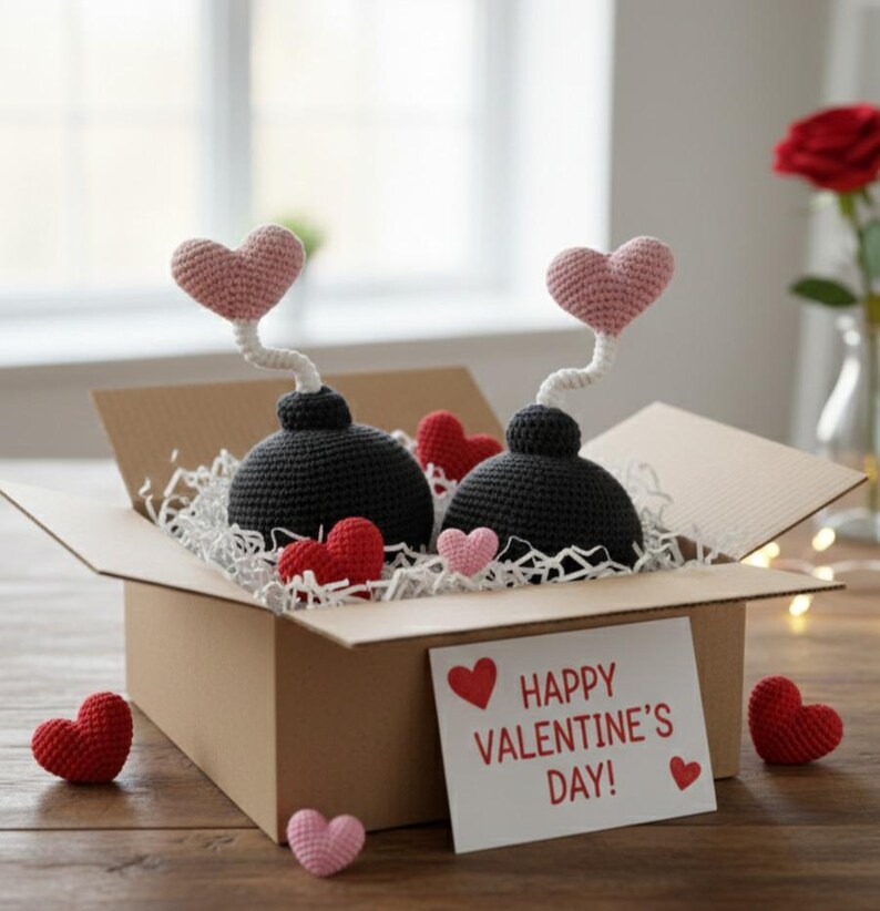 May include: A cardboard box filled with crochet Valentine's Day decorations. Two black crochet bombs with pink heart tops sit among red and pink crochet hearts. A card reads "Happy Valentine's Day!"