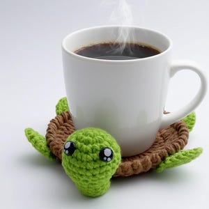 May include: A white ceramic mug filled with steaming black coffee rests on a handmade crochet turtle coaster. The coaster features a green turtle head with black eyes and a brown shell with green flippers.