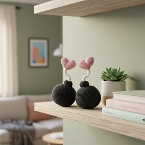 May include: Two handmade crochet decorative items shaped like bombs with pink heart tops. The bombs are dark gray, and the hearts are light pink. They sit on a wooden shelf with a succulent in a white pot and books.