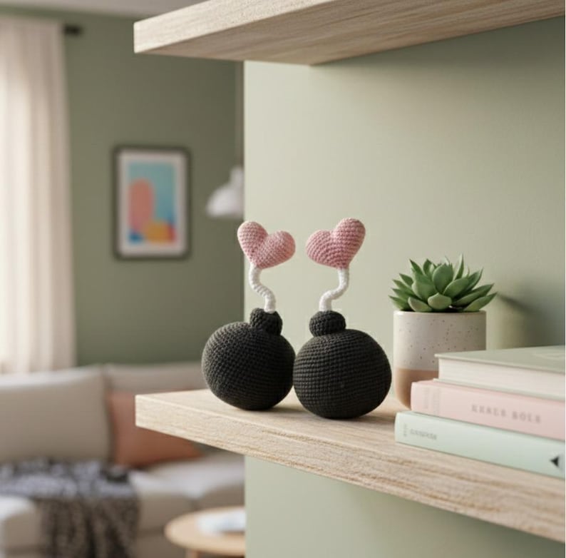 May include: Two handmade crochet decorative items shaped like bombs with pink heart tops. The bombs are dark gray, and the hearts are light pink. They sit on a wooden shelf with a succulent in a white pot and books.