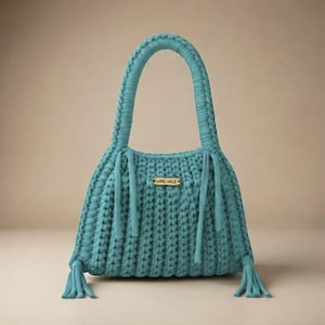 May include: A teal crochet handbag featuring a thick braided handle, a rectangular gold-toned metal tag with the text HAND MADE, and decorative tassels hanging from the bottom corners and the base of the handle on both sides of the bag.