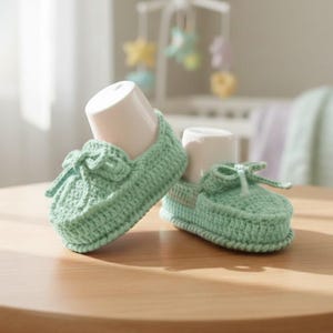 Puede incluir: Un par de patucos de bebé de ganchillo verde menta con un detalle de lazo. Los patucos se exhiben sobre una superficie de madera, con un fondo suave que incluye un móvil y una cuna para bebés.