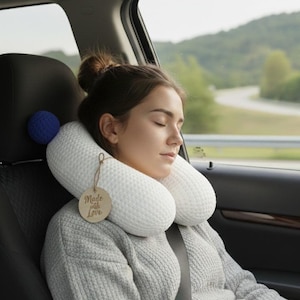Może przedstawiać: Biała dzianinowa poduszka podróżna z niebieskim pomponem przymocowanym do fotela samochodowego. Poduszka jest wokół szyi osoby ubranej w szary sweter. Na poduszce znajduje się metka z napisem "Made with Love."