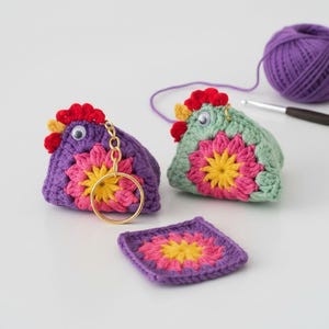 Peut inclure: Deux porte-clés poulet crochetés à la main, un violet et un vert, avec un anneau doré. Chaque poulet a une crête rouge, un bec jaune et un motif floral rose et jaune. Un petit carré crocheté et une pelote de laine violette sont également présents.