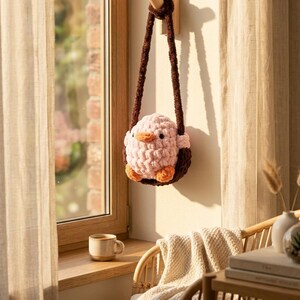 May include: A handmade, pink and brown crochet bird-shaped bag with a brown strap, hanging on a wooden peg. The bird has orange feet and beak. The bag is hanging near a window with a cup of coffee on the windowsill.