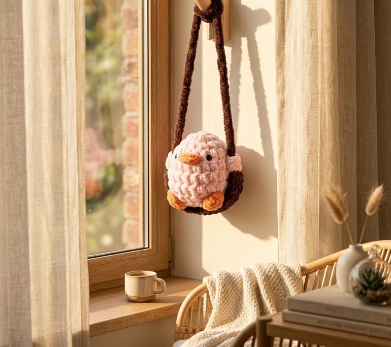 May include: A handmade, pink and brown crochet bird-shaped bag with a brown strap, hanging on a wooden peg. The bird has orange feet and beak. The bag is hanging near a window with a cup of coffee on the windowsill.