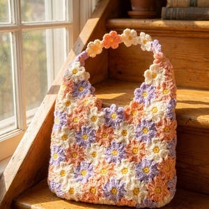 May include: A handmade crochet tote bag featuring a daisy pattern in shades of peach, lavender, cream, and yellow. The bag has a scalloped top edge and a handle made of crocheted flowers. The bag is sitting on wooden stairs.