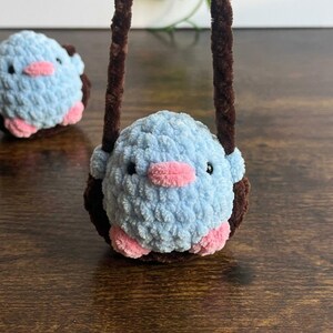 May include: A handmade, light blue crocheted bird with pink accents, including a beak and feet. The bird is inside a brown crocheted nest with a brown handle. Another bird is visible in the background.
