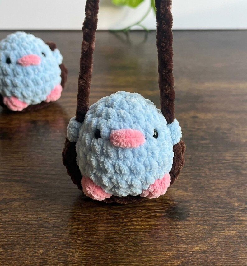 May include: A handmade, light blue crocheted bird with pink accents, including a beak and feet. The bird is inside a brown crocheted nest with a brown handle. Another bird is visible in the background.