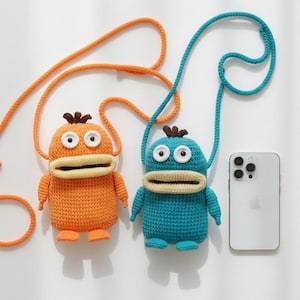 Puede incluir: Dos bolsos de crochet hechos a mano con forma de personajes de dibujos animados. Uno es naranja con la parte superior marrón y el otro azul con la parte superior azul. Ambos tienen ojos blancos, bocas beige y correas largas. Un smartphone blanco está al lado de los bolsos.
