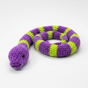 May include: A coiled plush snake toy. The snake has a purple head and body with alternating bands of lime green and purple. The toy has small black eyes and is made of a soft, textured material.